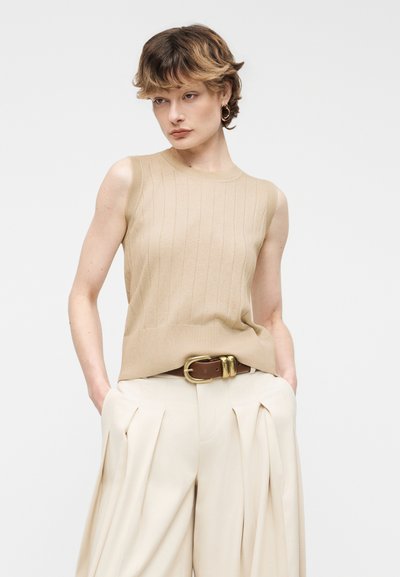 Άτομο με κοντά κυματιστά μαλλιά που φοράει μπεζ πλεκτό τοπ χωρίς μανίκια, κρεμ πλισέ παντελόνι με φαρδιά πόδια, καφέ ζώνη με χρυσή αγκράφα, με τα χέρια στις τσέπες.