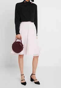 Blusa negra de manga larga con botones, falda A-line rosa claro, tacones negros con tiras, y bolso redondo burdeos con acentos dorados.