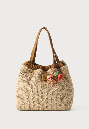 Sac cabas tissé beige avec bordure en tissu marron, bretelles d’épaule et pompon coloré ainsi qu’un charm fleur attaché près du fermoir.