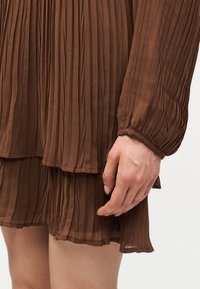 Χέρι δίπλα σε καφέ πλισέ φούστα και μακρυμάνικη μπλούζα με ελαστική μανσέτα, που δείχνει υφή ελαφριάς ύφανσης και λεπτομέρειες με στρωμένο τελείωμα.