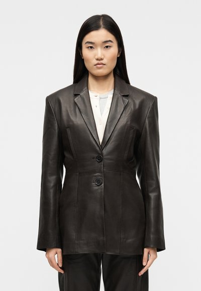 Femme aux longs cheveux noirs portant un blazer en cuir marron foncé ajusté sur un haut crème, debout devant un fond clair uni, face à l'avant.