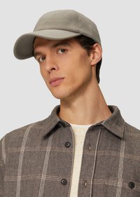 Grijze stoffen pet met een gebogen klep, gedragen door een model in een donker geruite flanellen overhemd over een lichtbeige trui. Textuurdetails zichtbaar.