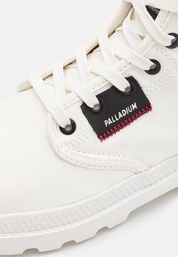 Bílé plátěné tenisky s černým štítkem s nápisem "Palladium", kovovými průvleky a bílými tkaničkami. Gumová podrážka s texturovaným vzorem pro lepší přilnavost.