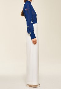 Blaues transparents Bluse mit floraler Stickerei an den Ärmeln, kombiniert mit hoch taillierten weißen weiten Hosen. Auffälliger Kragen und geknöpfte Manschetten.