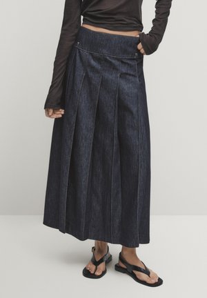 Femme portant une jupe midi plissée en jean foncé, un haut noir à manches longues et des sandales plates noires, debout devant un fond clair uni.