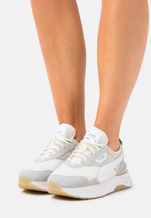 Hvide og grå Puma-sneakers med beige såler båret på bare fødder mod en ensfarvet lys baggrund.