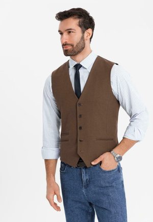 Uomo con capelli corti e scuri e barba, che indossa un gilet marrone abbottonato sopra una camicia a righe con maniche risvoltate, jeans blu e un orologio da polso.