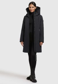 Sort vandtæt jakke med høj krave og lynlås, der har et mærke og lange ærmer, båret over en sort turtleneck og leggings.