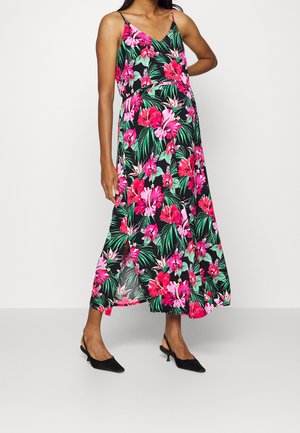 Femme portant une robe maxi noire sans manches avec un imprimé floral rose vif et vert et des talons slingback noirs, debout avec une main sur la hanche.
