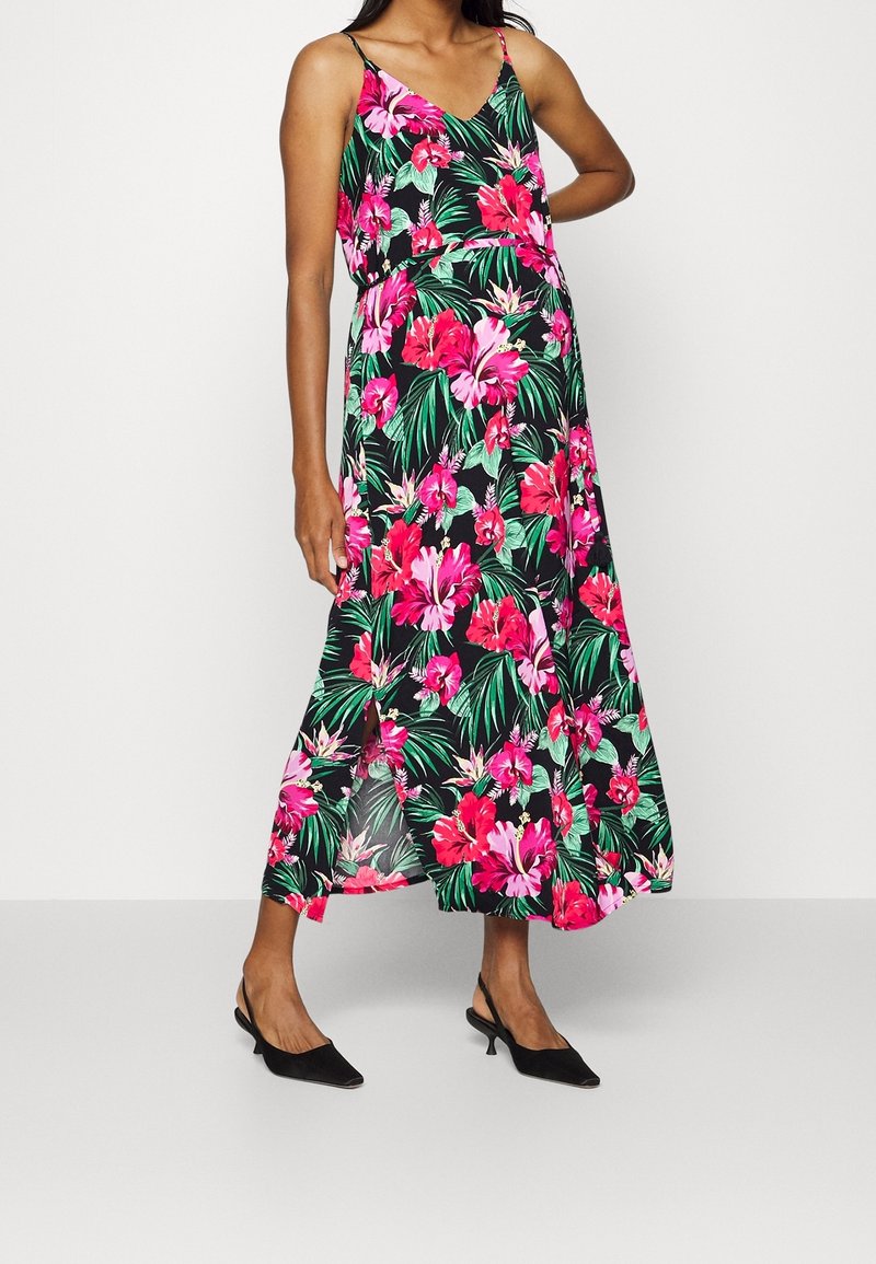 Femme portant une robe maxi noire sans manches avec un imprimé floral rose vif et vert et des talons slingback noirs, debout avec une main sur la hanche.