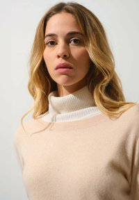 Maglione beige con un morbido colletto alto bianco a costine. Il tessuto appare liscio e il design è attillato con una sottile texture.