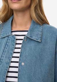 Světle modrá džínová bunda se stříbrnými patentními knoflíky a klasickým límcem. V kombinaci s černobílým pruhovaným tričkem pod ní.