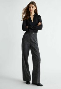 Blusa nera a maniche lunghe con scollo a V e dettaglio in catena dorata, abbinata a pantaloni grigi a gamba larga e vita alta con texture e calzature nere.