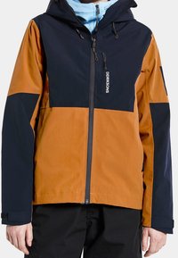 Color-blocked jacket featuring navy blue and brown sections, made from a durable material with a zippered front, adjustable hood, and cuffs.
