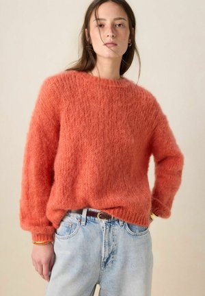 Femme portant un pull orange duveteux et un jean bleu clair avec une ceinture marron, debout avec une main dans la poche devant un fond uni.