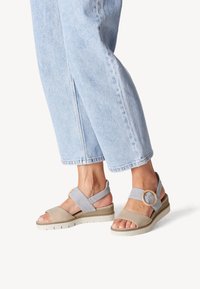 Sandales compensées grises et beiges avec une large lanière au niveau des orteils et des lanières croisées, ornées d'une boucle en bois, assorties à un jean bleu clair.