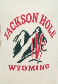 Sweter z dzianiny z napisem "JACKSON HOLE" i "WYOMING" w kolorze czerwonym, z graficznym motywem gór i narciarstwa w kolorze zielonym na kremowym tle.