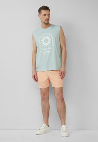 Helles blaues ärmelloses Shirt mit weißem Blumenmotiv, kombiniert mit korallfarbenen Shorts. Model trägt weiße Sneakers vor einem neutralen Hintergrund.