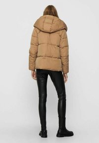 Piumino marrone chiaro con cappuccio ampio, texture trapuntata e cuciture frontali. Abbinato a pantaloni aderenti neri e stivali bassi neri.