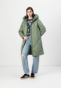 Manteau long matelassé vert avec capuche, fermeture éclair et poches latérales, porté sur un t-shirt graphique et un jean bleu déchiré.
