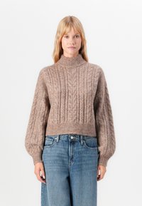 Kobieta ubrana w beżowy sweter w splot warkoczowy z bufiastymi rękawami i niebieskie dżinsy o wysokiej talii, stojąca na białym tle.