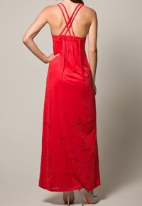 Vestido largo rojo con escote halter y tirantes entrelazados, que presenta una textura suave y patrones decorativos de hojas.