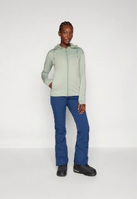 Svetlo zelena jopa z zadrgo iz teksturirane tkanine, v kombinaciji z modrimi smučarskimi hlačami in črnimi škornji. Ponaša se z ruto in stranskimi žepi.