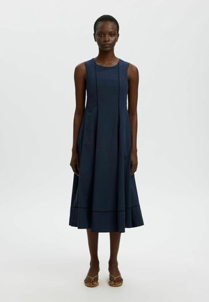 Femme portant une robe midi bleu marine sans manches avec un corsage cintré et une jupe évasée, associée à des sandales couleur beige, debout devant un fond uni.