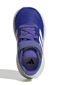 Zapatilla deportiva morada con parte superior de malla, rayas blancas, correa ajustable, lengüeta acolchada y suela texturizada. Detalles en verde lima en el talón.