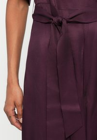 Detalhe de um vestido de cetim roxo, com um nó na cintura, textura suave e um design elegante e fluido com costuras verticais.