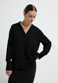 Black button-up shirt with a subtle leopard print, featuring long sleeves and a relaxed fit. Worn with a silver neckpiece. Simple, elegant design.