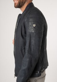Giacca in pelle blu navy con spalle testurizzate, chiusura con zip e dettaglio a scatto in vita. Presenta una medaglia con logo sulla manica sinistra.