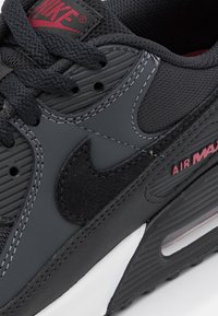 Sneaker Nike Air Max avec une tige en synthétique noir, des panneaux texturés, des accents en daim noir et une semelle intermédiaire blanche avec des détails de logo rouges.