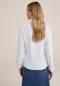 Blusa blanca de manga larga con dobladillo redondeado y puños abotonados, combinada con una falda de mezclilla azul. Tela suave con un corte clásico.