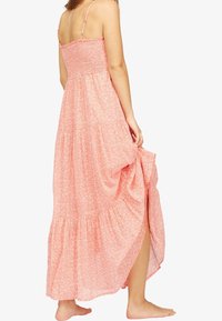 Robe maxi longue à fleurs roses avec des bretelles fines, un corsage smocké, une jupe à volants et une fente sur le côté pour faciliter les mouvements. Tissu doux et léger.
