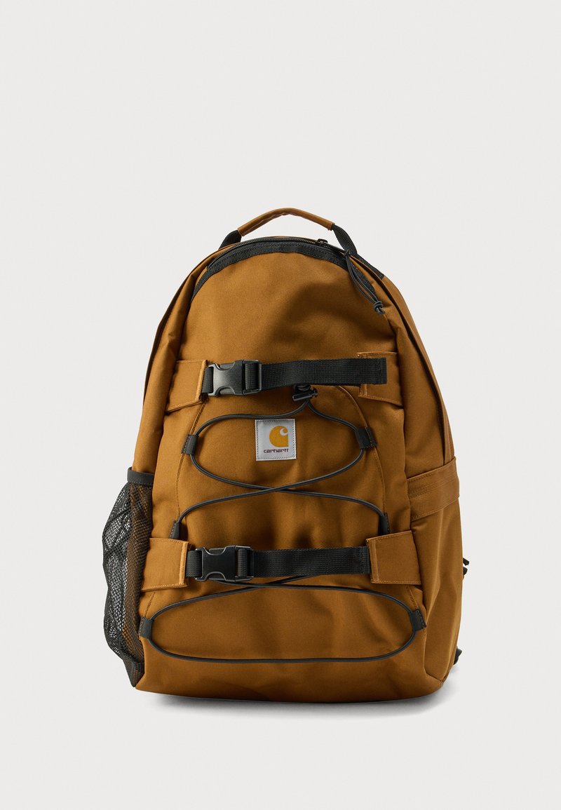 Sac à dos marron avec bretelles réglables, boucles avant et une poche latérale en filet. Comprend un patch logo, un tissu résistant et un détail de cordon élastique.