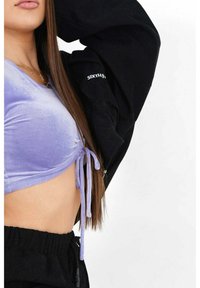 Vrouw in een lila gerimpelde crop top met strikjes en zwarte trainingsbroek, met een zwarte jas die gedeeltelijk over haar schouder hangt.