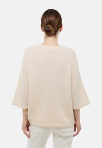 Femme aux cheveux bruns en chignon, portant un pull beige en tricot lâche à manches trois-quarts et un pantalon blanc, se tenant devant un fond uni.