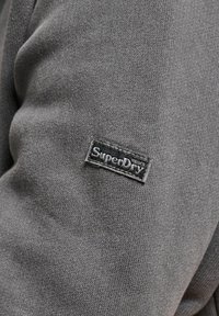 Material de pulover gri cu o textură moale și un design ajustat. Prezintă un patch cu logo-ul negru "SuperDry" pe mâneca stângă.