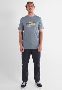 T-shirt en coton bleu clair avec un imprimé graphique d'un coucher de soleil et de vagues, associé à un pantalon navy foncé et des chaussures de skate noires avec des lacets blancs.