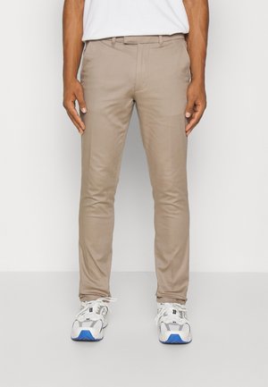 Man draagt beige slim-fit broek en witte sneakers met blauwe zolen, staand tegen een effen achtergrond.