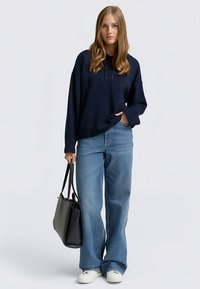 Tengerészkék oversized kapucnis pulóver világoskék, bő szárú farmerral és fehér sportcipővel párosítva. Fekete kézitáska, laza szabású és casual stílusú.