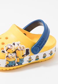 Gul träskor med blå hälrem, prydda med Minion-karaktärer på sidan. Tillverkade av plast med ventilationshål, slät yta och tryckta grafik.