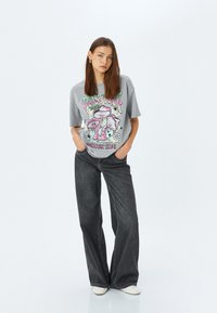 Camiseta gris de gran tamaño con un diseño gráfico colorido de hongos y flores, combinada con jeans negros sueltos y zapatillas blancas.
