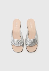 Silverfärgade slip-on sandaler med en vriden remdesign och en slät, naken sula. Sulan är platt med en lätt struktur för grepp.
