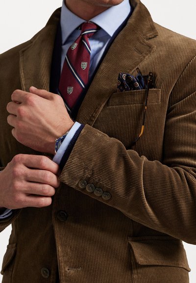 Blazer en velours côtelé marron avec revers en pointe, doté de poches latérales, d'une chemise bleue en dessous et d'un mouchoir de poche à motifs.
