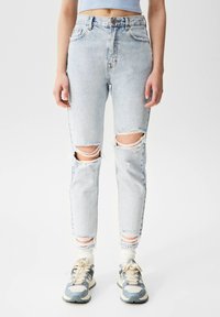 Jeans desgastados de color azul claro con rodillas rasgadas, presentan una cintura alta y un corte ajustado. La tela tiene una textura suave y usada, combinada con zapatillas gris y blancas.