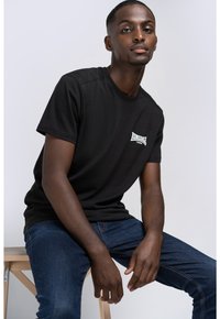 Sort bomulds t-shirt med korte ærmer og rund halsudskæring, med et hvidt logo på venstre bryst. Modellen sidder ned og har blå jeans på.