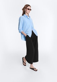 Femme en chemise bleue claire à boutons, pantalon noir à jambes larges, tongs noires, foulard marron à motifs et lunettes de soleil, debout les mains dans les poches.