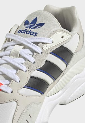 Bílo-béžové tenisky Adidas s černými a modrými pruhy, bílými tkaničkami a polstrovaným límcem, zobrazující logo Adidas na jazyku.